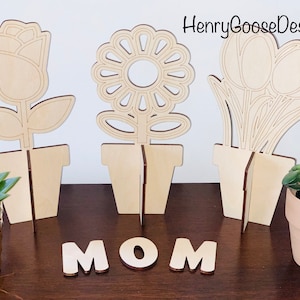 May include: Three wooden flower pot decorations with a rose, sunflower, and tulip design. The pots are standing on a wooden surface with the word "MOM" spelled out in wooden letters.