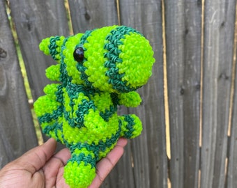 May include: A handmade, green and dark green striped dragon figurine. The dragon is made of small rubber bands, with a black eye. The dragon is held in a hand against a wooden fence background.