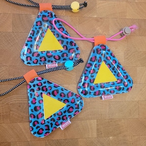May include: Three triangular reflective safety tags featuring a blue and pink leopard print pattern with a central yellow triangle. Each tag has an orange fabric loop, a small white tag labelled BLUESUG, and an adjustable cord with plastic beads.