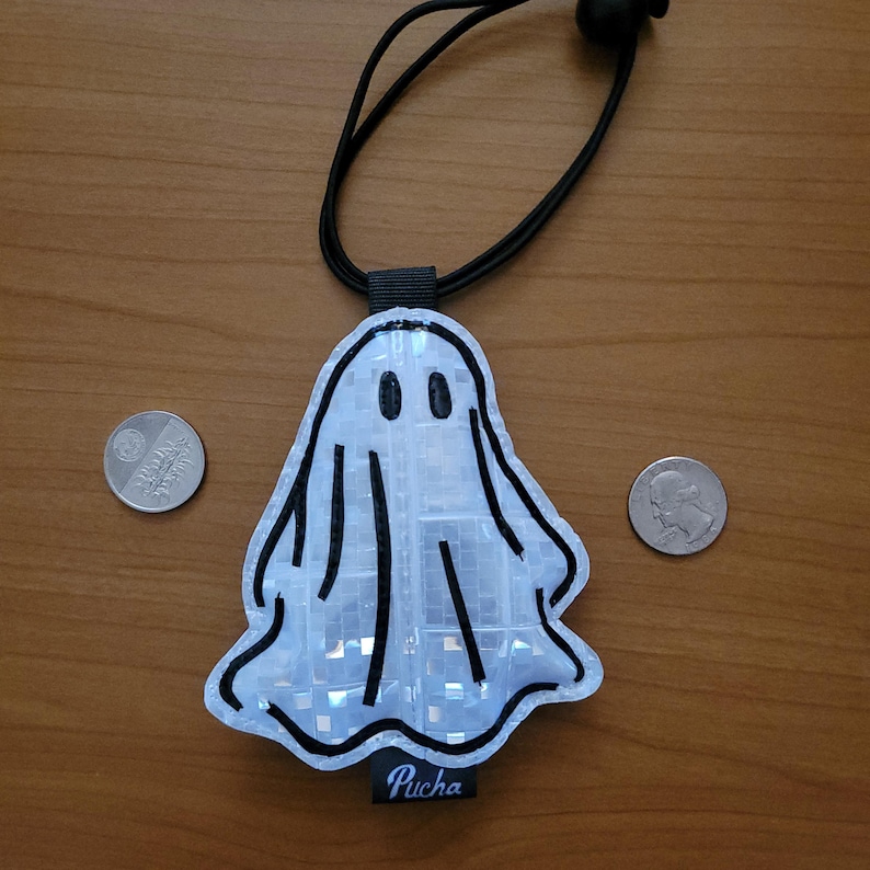 May include: A white ghost-shaped reflective charm with black outlines and details, suspended from a black cord. The charm features two black oval eyes and vertical lines. The word "Pucha" is printed on a black tag at the bottom.