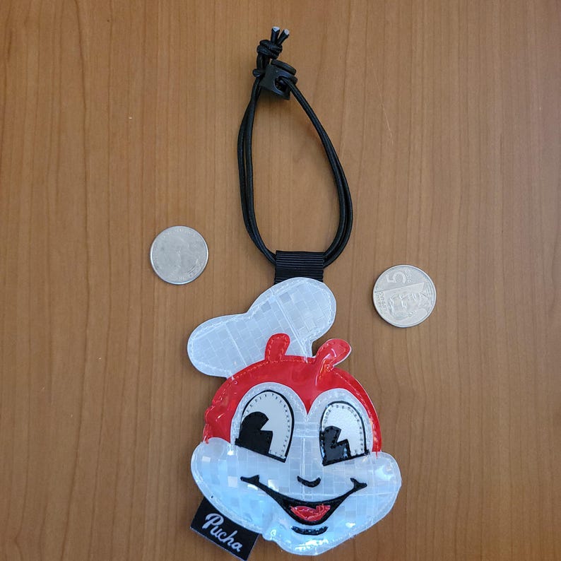 May include: A red and white reflective pendant with a cartoon character face, a black cord, and a clasp. The pendant has the word "Pucha" on a small black tag. Two coins are next to the pendant.