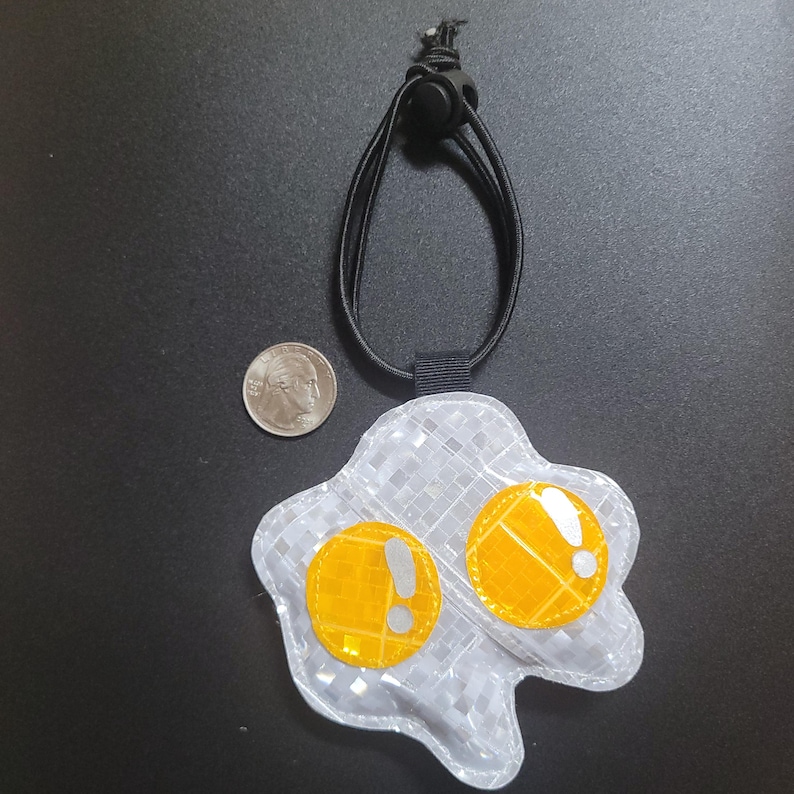 May include: A reflective, egg-shaped pendant with two yellow yolks and a grey, chequered white base. The pendant has a black cord and an adjustable toggle. A coin is next to the pendant for size comparison.