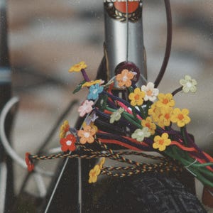 Peut inclure: Une collection de tiges de fleurs crochetées colorées attachées à un cadre de vélo argenté avec un logo Kuwahara. Les tiges sont faites de cordons tressés de différentes couleurs, chacune surmontée de petites fleurs plates à cinq pétales.