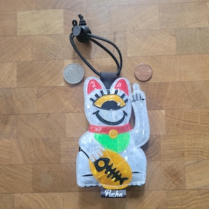 May include: A reflective white lucky cat charm with red, green, and yellow details. The cat is giving the middle finger and holding a fish. The charm has a black cord and the word "Pucha" on the bottom.