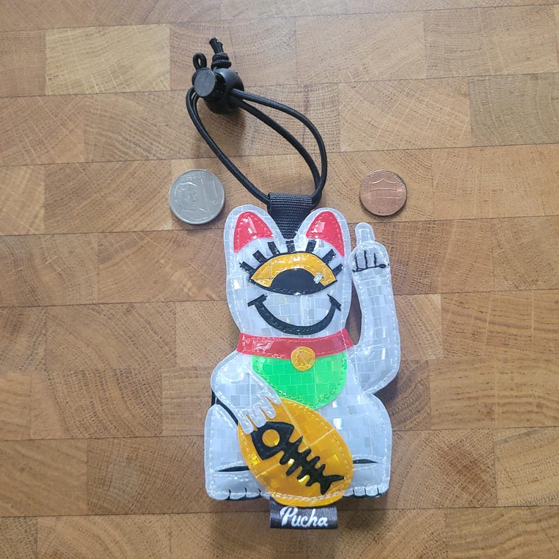 May include: A reflective white lucky cat charm with red, green, and yellow details. The cat is giving the middle finger and holding a fish. The charm has a black cord and the word "Pucha" on the bottom.