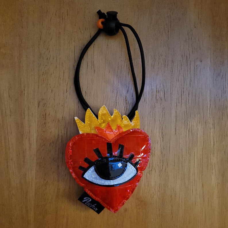 May include: A red heart-shaped pendant with a stylized eye design, topped with a yellow crown. The pendant hangs from a black cord with an adjustable toggle. The word "Pucha" is printed on a small black tag attached to the pendant.