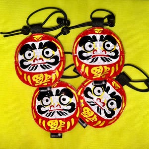 May include: Four circular Daruma doll patches in red, white, and black with gold accents. Each patch features a stylised face with large eyes, a moustache, and Japanese kanji characters. They include black elastic cords with toggles.