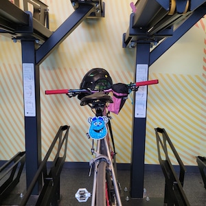 May include: A bicycle with a black helmet, red handlebar grips, and a blue monster-shaped tag is parked in a bike rack. The bike has a black seat and a pink bag attached to the handlebars. The rack is black and mounted on a wall with yellow and white stripes.