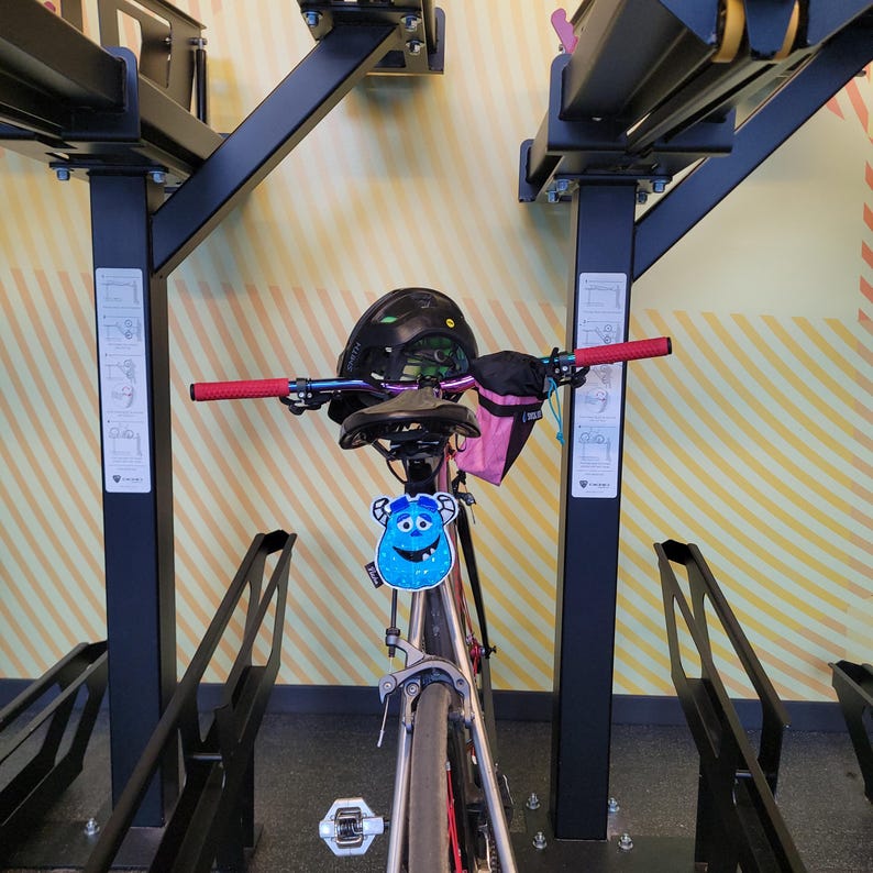 May include: A bicycle with a black helmet, red handlebar grips, and a blue monster-shaped tag is parked in a bike rack. The bike has a black seat and a pink bag attached to the handlebars. The rack is black and mounted on a wall with yellow and white stripes.