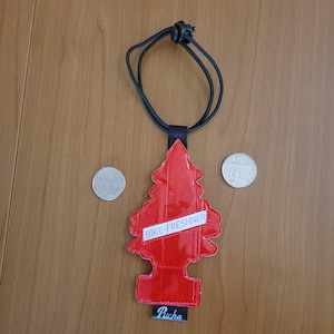 May include: A red, tree-shaped air freshener with the text "BIKE-FRESHNER" on a white banner. The air freshener hangs from a black cord. The word "Pucha" is printed at the bottom. Two coins are next to the air freshener.