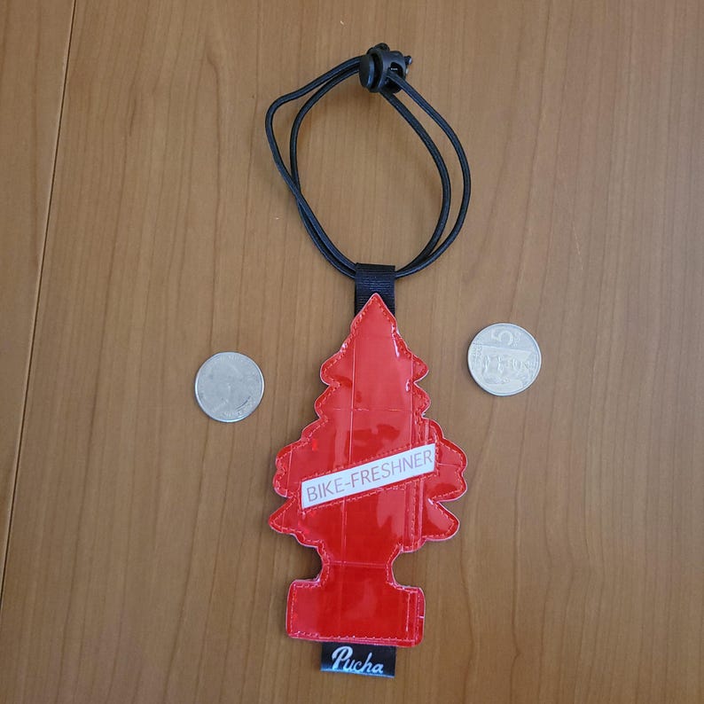 May include: A red, tree-shaped air freshener with the text "BIKE-FRESHNER" on a white banner. The air freshener hangs from a black cord. The word "Pucha" is printed at the bottom. Two coins are next to the air freshener.