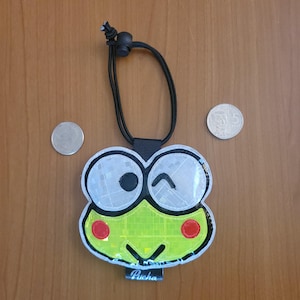 May include: A reflective frog-shaped charm with a black elastic cord and toggle. The charm features large white eyes, a lime green face, red circular cheeks, and a black mouth. The word Pucha is printed on a small black tag at the bottom.