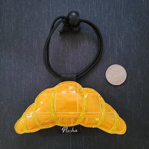 May include: A yellow croissant-shaped charm with a black elastic band and a black bead. The charm has a grid-like pattern and is accented with yellow trim. A coin is next to the charm. The charm has the word "Pucha" on it.