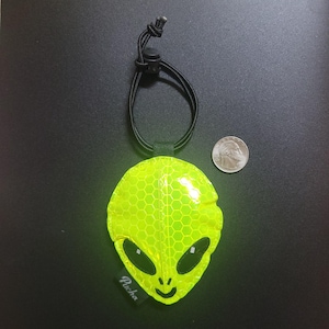 May include: A fluorescent yellow alien-shaped reflective tag with a black cord and adjuster. The tag features a honeycomb pattern, black eyes, and a mouth. A coin is next to the tag for size comparison. The tag has the word "Pucha" printed on it.