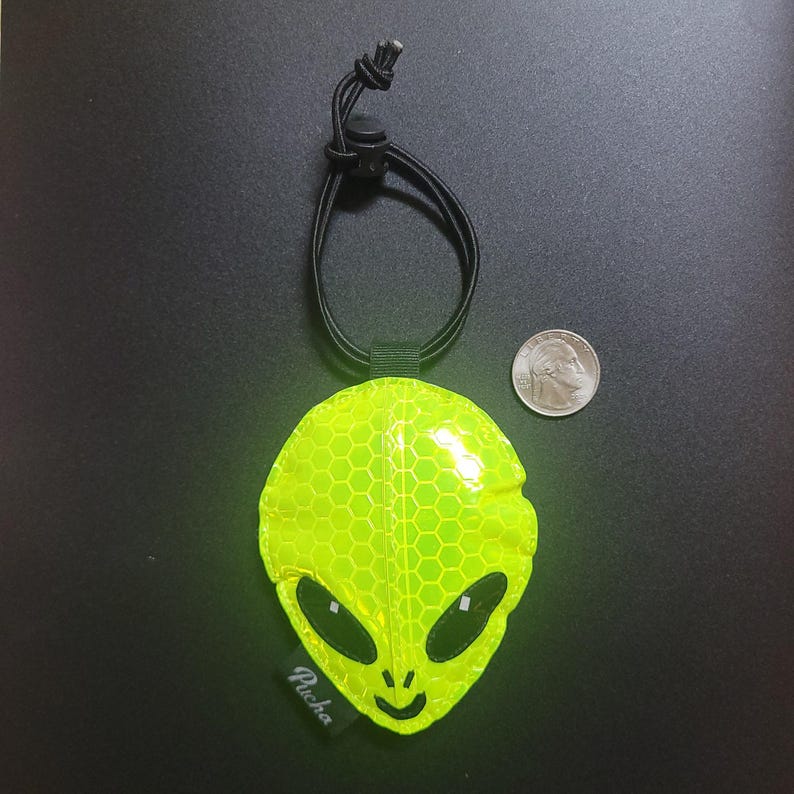 May include: A fluorescent yellow alien-shaped reflective tag with a black cord and adjuster. The tag features a honeycomb pattern, black eyes, and a mouth. A coin is next to the tag for size comparison. The tag has the word "Pucha" printed on it.