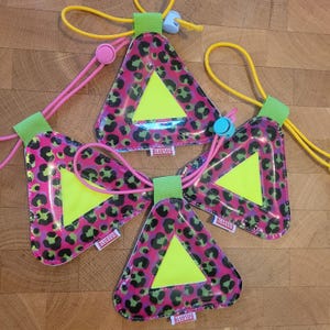 Puede incluir: Tres etiquetas de seguridad reflectantes triangulares con un estampado de leopardo en rosa, verde y negro. Cada etiqueta tiene un centro triangular amarillo brillante, un lazo de tela verde claro en la parte superior y un cordón amarillo o rosa con un tope de plástico. El texto BLUESUG está impreso en una pequeña etiqueta blanca unida al lateral de cada artículo.