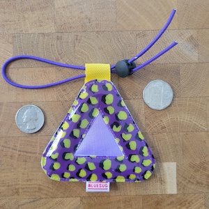 Puede incluir: Una etiqueta reflectante triangular con estampado de leopardo en morado y verde lima, con un lazo de tela amarillo, un cordón elástico morado con un cierre negro y una pequeña etiqueta blanca con el texto BLUESUG, entre dos monedas.