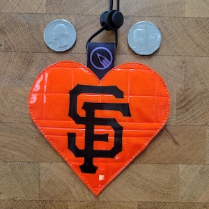 Puede incluir: Una etiqueta reflectante naranja en forma de corazón con el logotipo negro de los San Francisco Giants en el centro. La etiqueta incluye un cordón negro con un cierre de plástico y una pequeña lengüeta negra con un icono de lápiz circular.