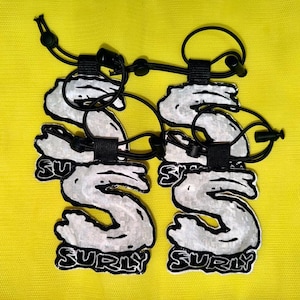 May include: Two reflective silver patches shaped like the letter S, each featuring the word SURLY in black lettering at the bottom. The patches are attached to black elastic cords with adjustable toggles, designed for secure attachment to gear or clothing.