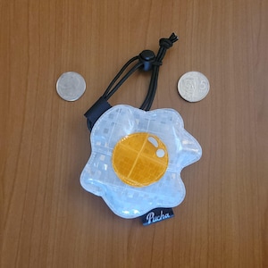 May include: A clear, egg-shaped keyring with a yellow yolk design and a black cord. The keyring has a black tag with the word "Pucha" on it. The background is a wooden surface.