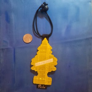 May include: A yellow, tree-shaped air freshener with the text "BIKE-FRESHNER" and "Pucha" on a black strap. The air freshener hangs from a black cord with a black bead. A coin is next to the air freshener.