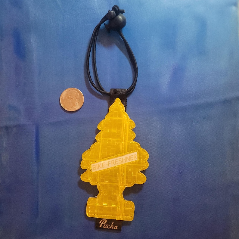 May include: A yellow, tree-shaped air freshener with the text "BIKE-FRESHNER" and "Pucha" on a black strap. The air freshener hangs from a black cord with a black bead. A coin is next to the air freshener.