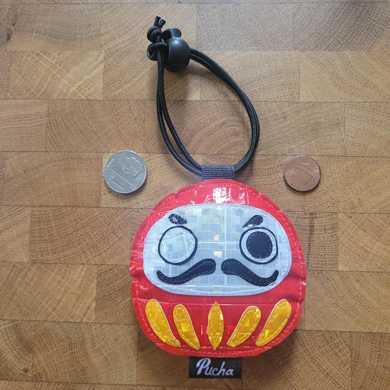May include: A red Daruma doll-shaped accessory with a black cord and toggle. The doll has a white face, black mustache, and yellow accents. The word "Pucha" is printed on the bottom. A 20 pence coin and a penny are next to the doll.