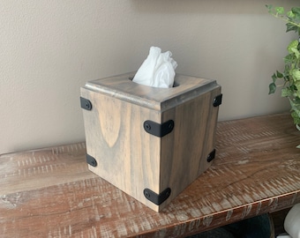 May include: A wooden tissue box cover with a gray wash finish and black metal accents. The box has a square shape and is sitting on a wooden surface.