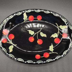 May include: A black oval serving dish with a white and green border and a design of red cherries with green leaves.
