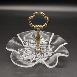 May include: A clear glass serving dish with a gold metal handle. The dish has a scalloped edge and a floral design.