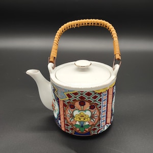 May include: A white ceramic teapot with a woven brown handle. The teapot has a colorful floral pattern with blue, red, yellow, and green accents.