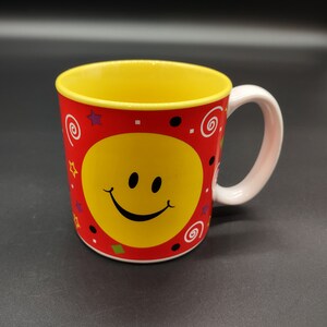 May include: A red ceramic mug with a yellow smiley face and colorful swirls and stars. The mug has a white handle.