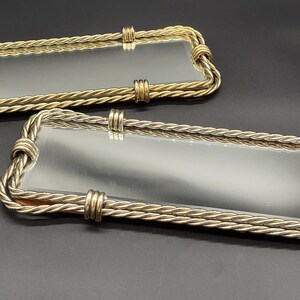 May include: Two rectangular decorative mirrors with gold rope-like frames. The mirrors are different sizes and are lying on a dark gray surface.