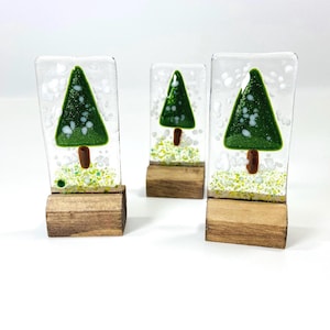 May include: Three decorative glass Christmas trees. Each tree has a green triangular design with white accents, a brown trunk, and sits on a wooden base. The background is white with a speckled design.