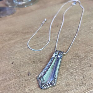 Può includere: Una collana con pendente in argento realizzata con posate riciclate. Il pendente ha un motivo floreale decorativo e una forma rettangolare. La catena della collana è sottile e argentata. Il pendente è lungo circa 5 cm.