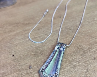 May include: A silver pendant necklace made from repurposed silverware. The pendant has a decorative floral design and a rectangular shape. The necklace chain is thin and silver. The pendant is approximately 2 inches long.