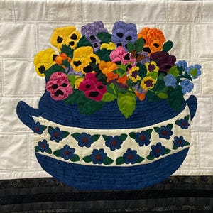 May include: A quilted wall hanging depicting a blue vase filled with vibrant pansies. The vase has a white band adorned with blue floral patterns. The backdrop is a white, textured fabric. The pansies are yellow, purple, pink, and orange.