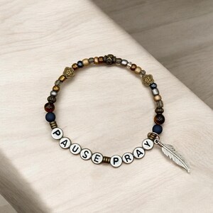 May include: A beaded bracelet with the words "PAUSE PRAY" spelled out in white letter beads. The bracelet features a variety of beads in gold, brown, and silver tones, with a silver feather charm. The bracelet is a spiritual or inspirational accessory.