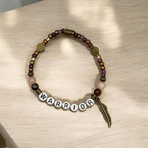 May include: A beaded bracelet with the word "WARRIOR" spelled out in white letter beads. The bracelet features a mix of purple, gold, and pink beads, with a decorative feather charm. The bracelet is on a light wood surface.