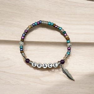 May include: A beaded bracelet with the word "GRACE" spelled out in white letter beads. The bracelet features a variety of colorful beads, including purple, blue, and gold, and a silver feather charm. The bracelet is on a light wood surface.