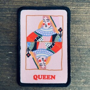 May include: A pink and black iron-on patch with a colorful illustration of a queen playing card. The queen is wearing a red, white, and blue dress and has a flower in her hand. The text "QUEEN" is printed in red at the bottom of the patch.