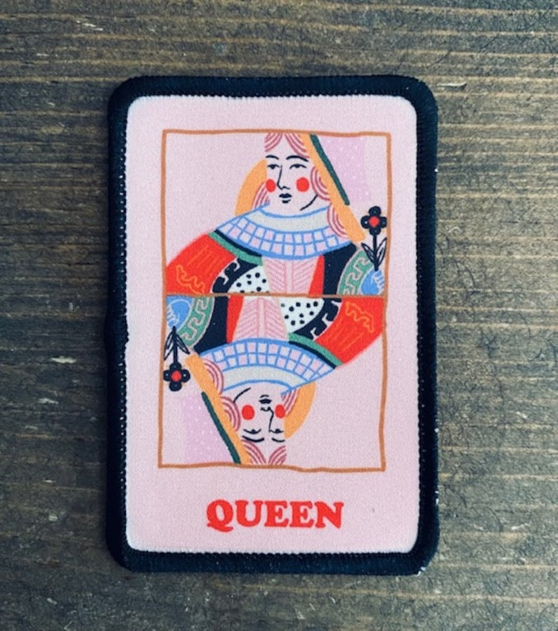 May include: A pink and black iron-on patch with a colorful illustration of a queen playing card. The queen is wearing a red, white, and blue dress and has a flower in her hand. The text "QUEEN" is printed in red at the bottom of the patch.