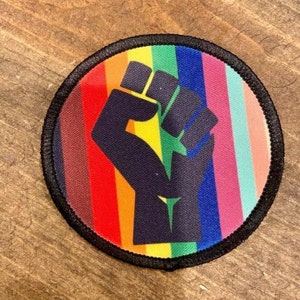 May include: A black fist raised in the air, with a rainbow background. The fist is a symbol of solidarity and resistance.