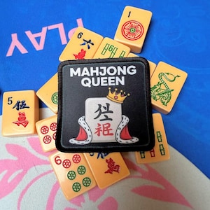 May include: A collection of ivory-colored Mahjong tiles scattered around a black patch. The patch reads "MAHJONG QUEEN" with a crown and a tile design. The tiles feature red, green, and black symbols and numbers.