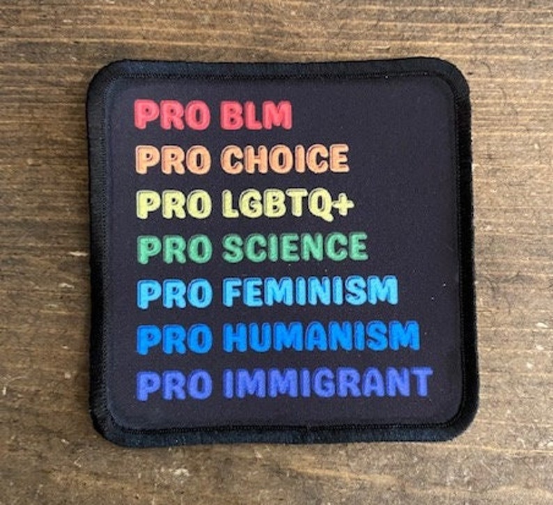 May include: A black square patch with white text in a rainbow color scheme. The text reads "PRO BLM", "PRO CHOICE", "PRO LGBTQ+", "PRO SCIENCE", "PRO FEMINISM", "PRO HUMANISM", and "PRO IMMIGRANT".