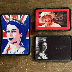 May include: Three black framed patches with images of Queen Elizabeth II. The first patch features a colorful image of the Queen with a crown and a Union Jack flag background. The second patch features a color image of the Queen wearing a hat and a red background with the text "Queen Elizabeth II 1926-2022". The third patch features a black and white image of the Queen wearing a crown and a black background with the text "Queen Elizabeth II 1926-2022".