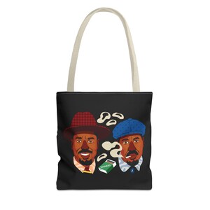 May include: Black tote bag featuring a colorful illustration of two men with hats, one wearing a red hat and the other a blue cap. The bag has a cream-colored handle and a pack of cigarettes is visible.