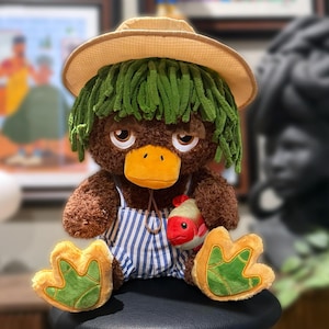 May include: A plush duck doll with brown fur, an orange beak, and green hair, wearing a straw hat and blue and white striped overalls. The doll holds a red fish and has large webbed feet. The doll is approximately 12 inches tall.