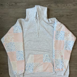 Puede incluir: Una sudadera con capucha gris con mangas y bolsillo delantero de patchwork. Las mangas y el bolsillo presentan un diseño de patchwork con estampados florales rosas, blancos y azules. La sudadera tiene un cordón y un bolsillo de canguro.