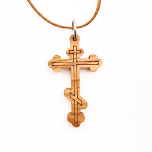 May include: Wooden Orthodox cross pendant with a light brown cord. The cross features a three-bar design and is made of light-colored wood with dark brown accents. The pendant hangs from a thin, tan-colored cord.